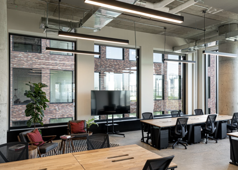 Open-plan office with natural light, modern furniture, and a lounge area near tall glass windows.